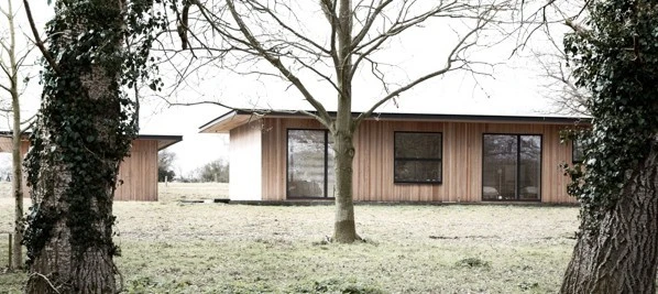 Moderní dřevěný bungalov v přírodě s velkými okny a okolním stromovým porostem.