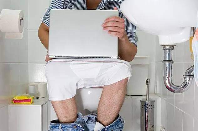 Muž sedí na záchodě s notebookem v koupelně.