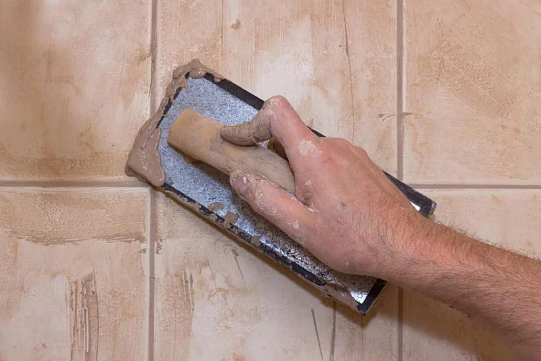 Ruční aplikace lepidla mezi dlaždicemi při renovaci koupelny.