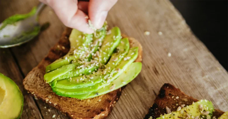 Ruce posypávají sezamové semínka na plátky avokáda na toastu.