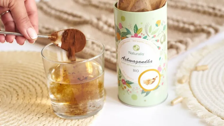 Ruka nalévá prášek ashwagandy do sklenice vedle balení.